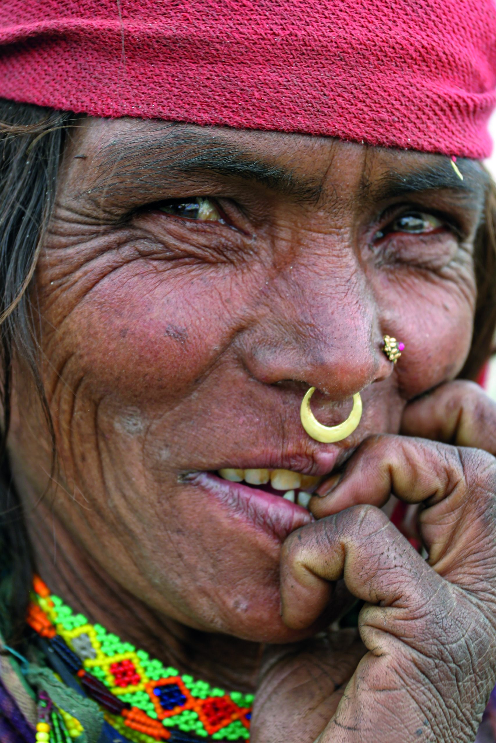 A friendly and curious woman's face, her fingers reaching for her mouth. The woman has a nose ring and age-related wrinkles around the eyes.