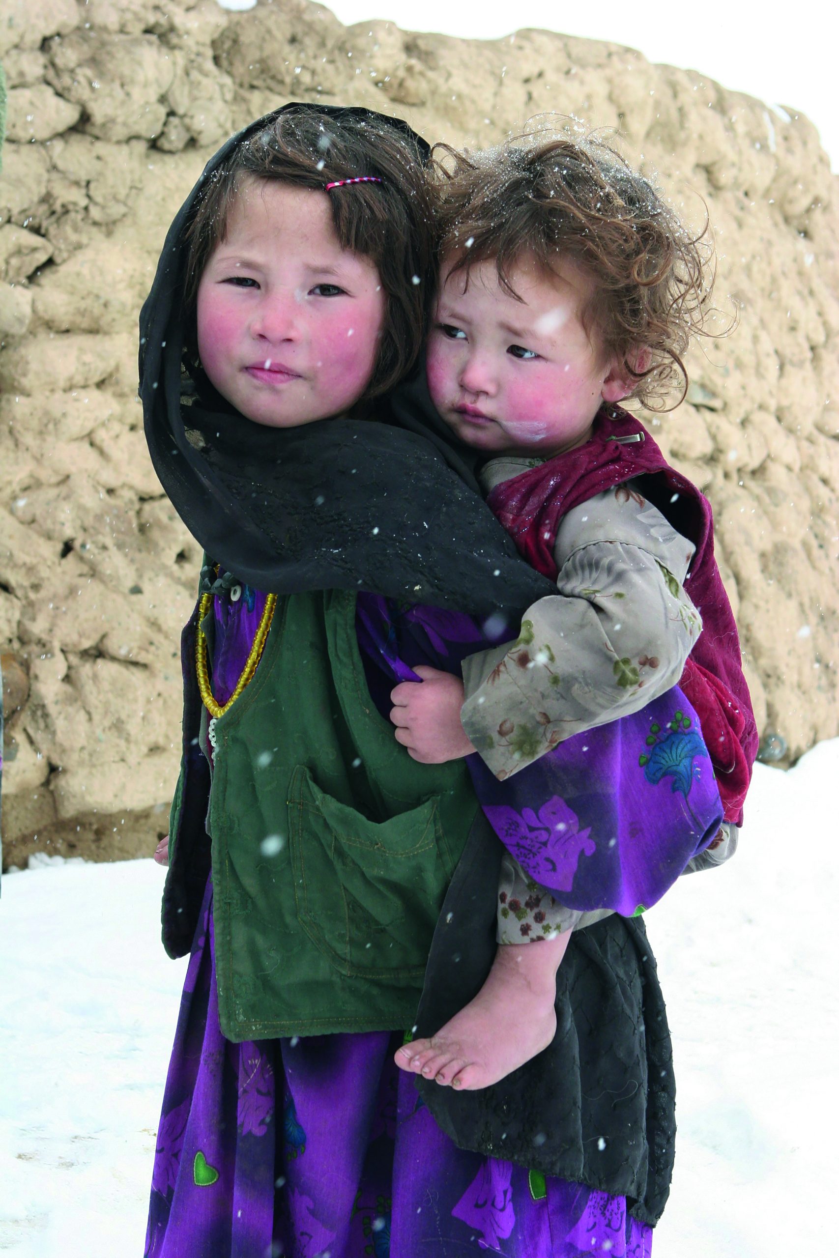 Big sister is carrying the smaller one on her back. She looks at the camera. Both have brown hair and red cheeks. Only the older one is wearing a scarf.