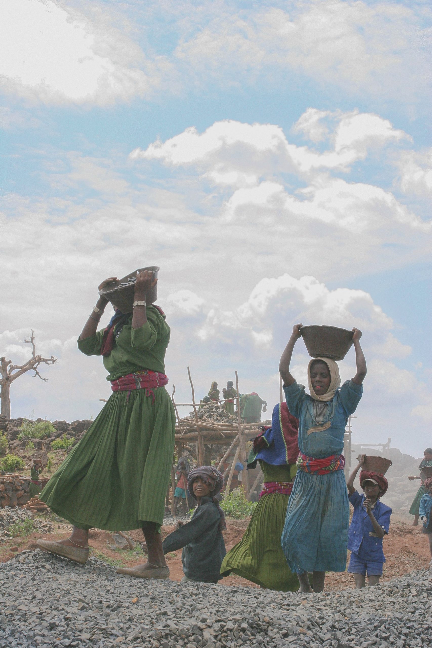 Women dressed in skirts carry water cans on their heads. Small children follow their mothers as they walk.