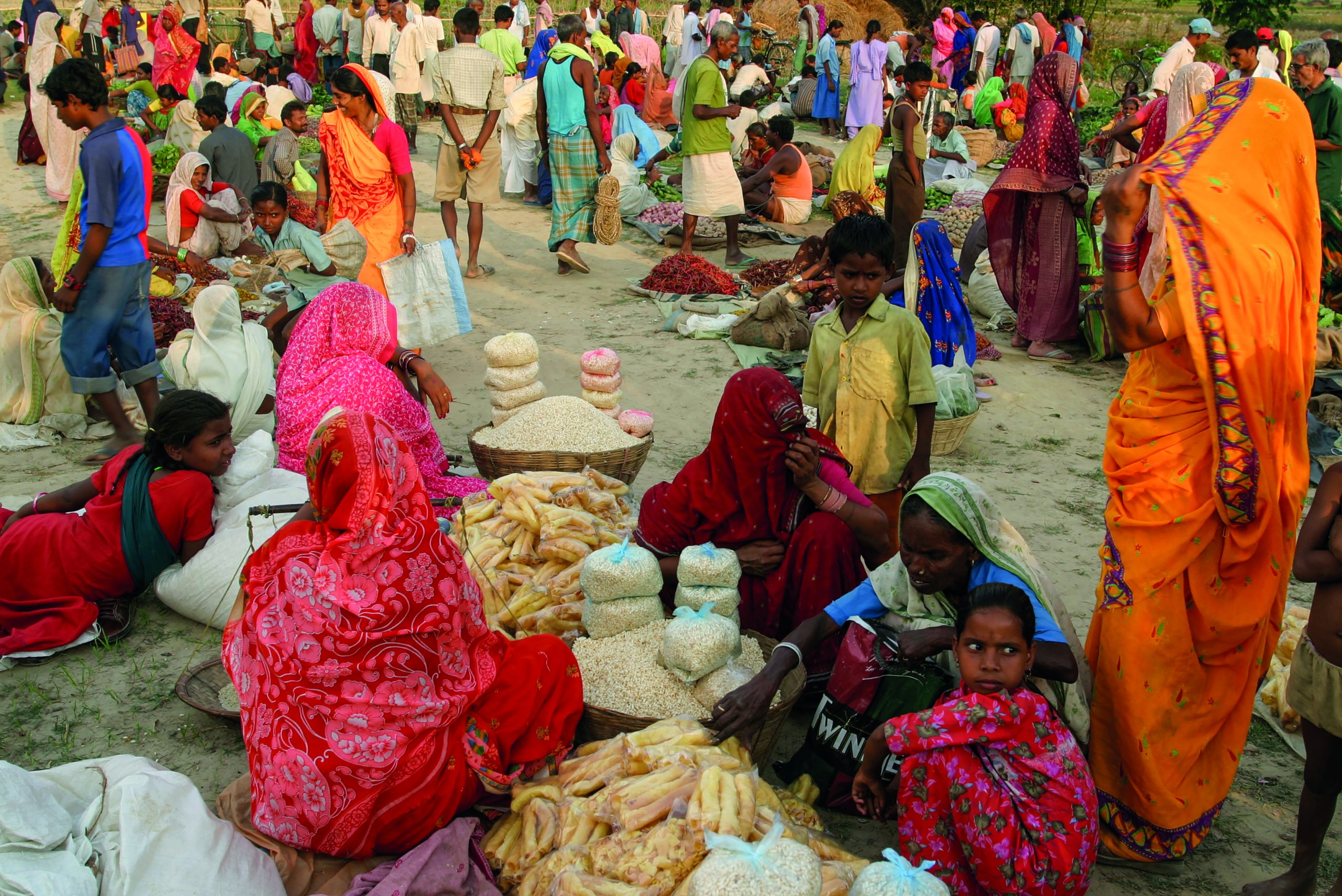 A lively view of the market. People are colourfully dressed and some are sitting on the ground. There are items for sale on the blankets, such as food.