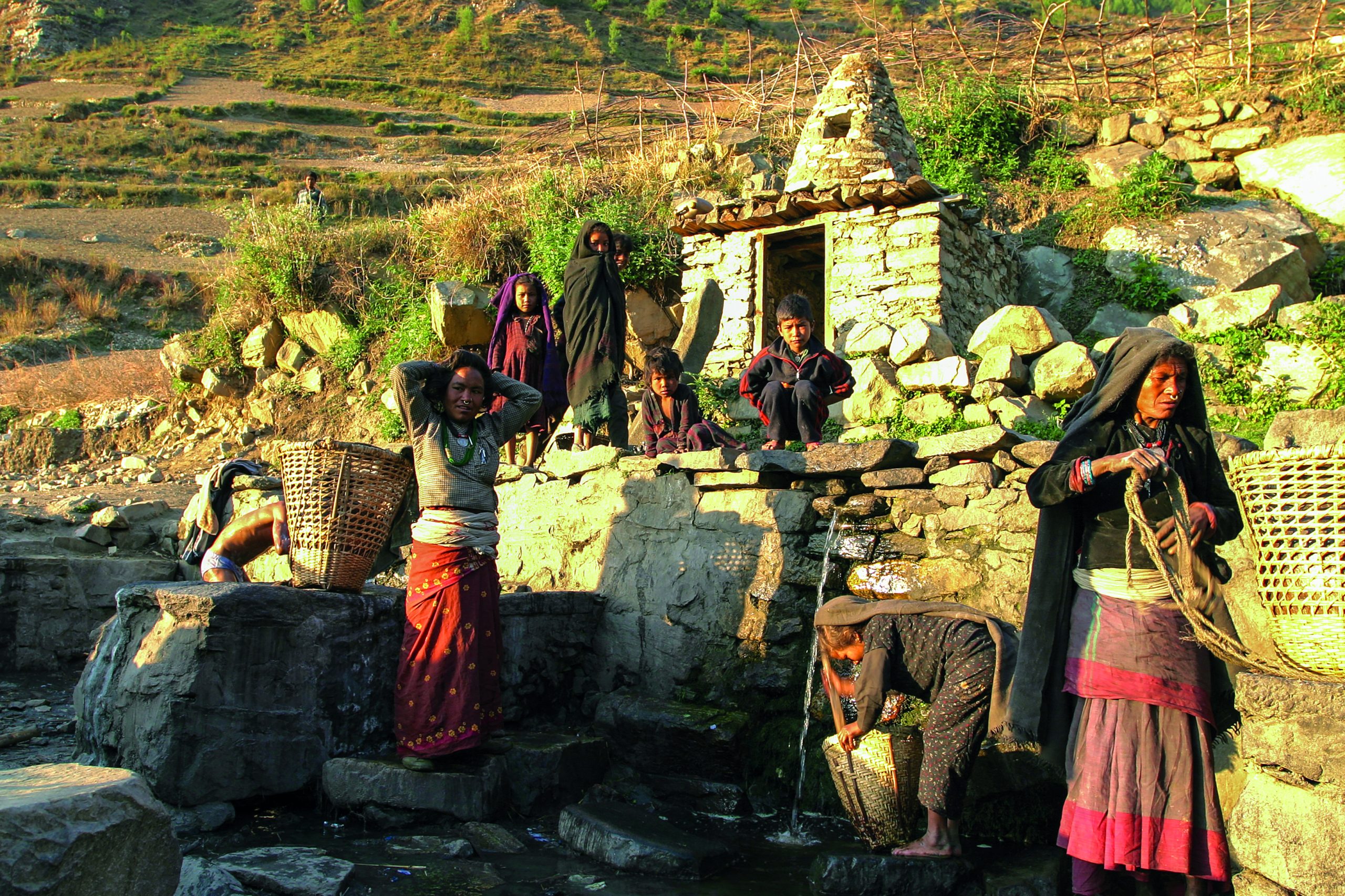Women carry water in baskets and children watch the action. The terrain is rocky. Water runs down a slope.