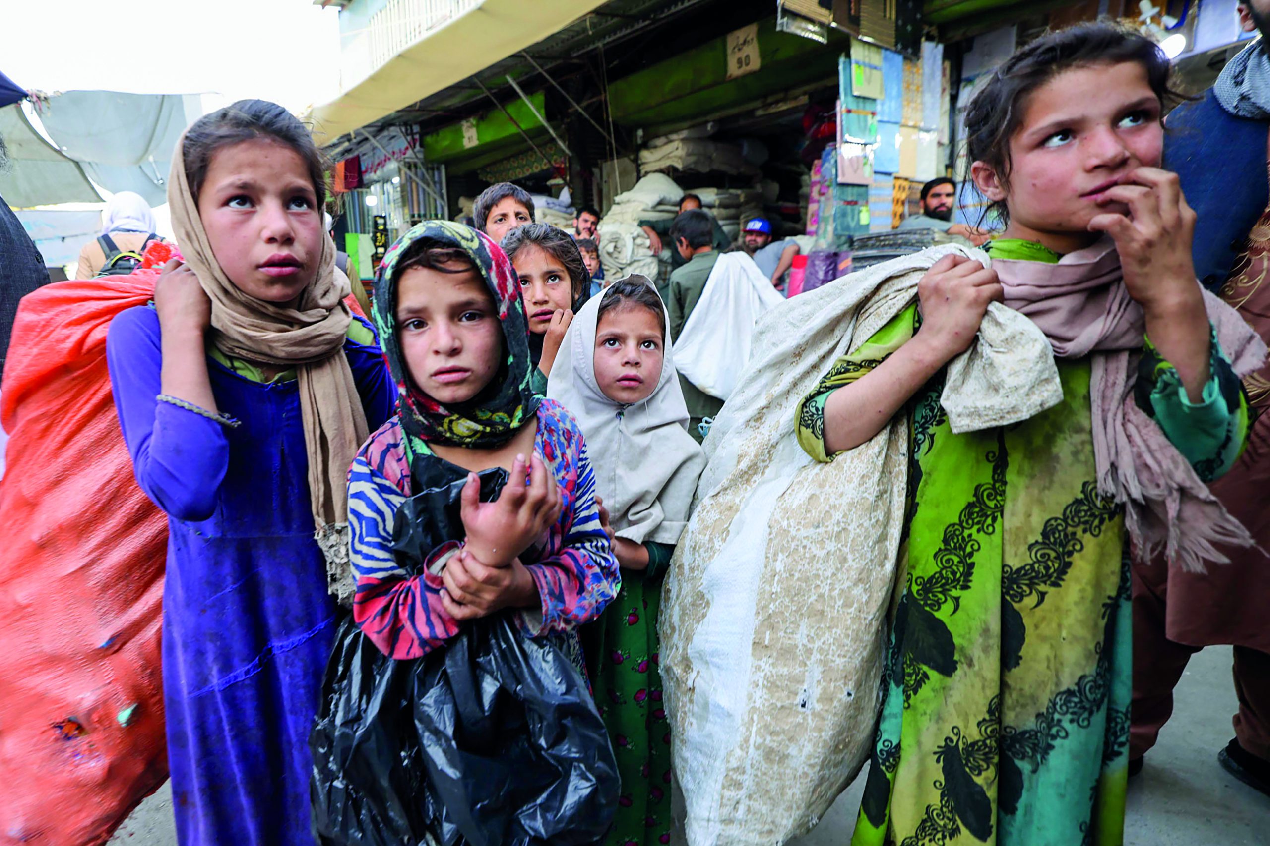 Neljä nuorta tyttöä tiiviissä ryhmässä markkinoilla. He kantavat tavaraa kangassäkeissä.