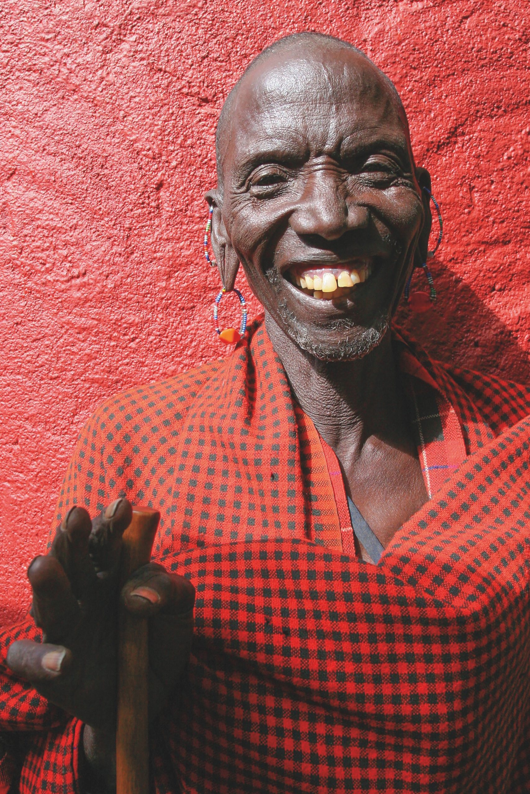 A dark-skinned man smiles. He is wearing earrings and a red and black checked shirt. The background is a red wall.