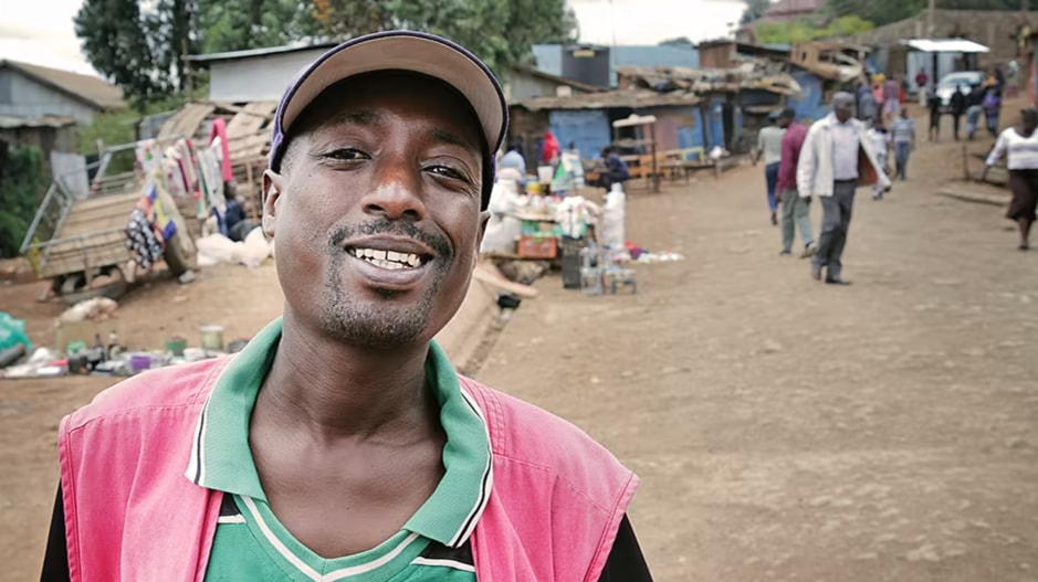 A man looks towards the camera. In the background you see shack buildings, carts and goods in piles.