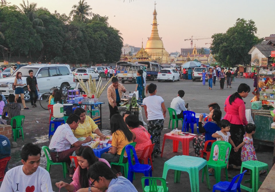A group of people are sitting around plastic tables in a café. There are a lot of people. In the background there are cars in the parking lot. A tower is visible on the horizon.